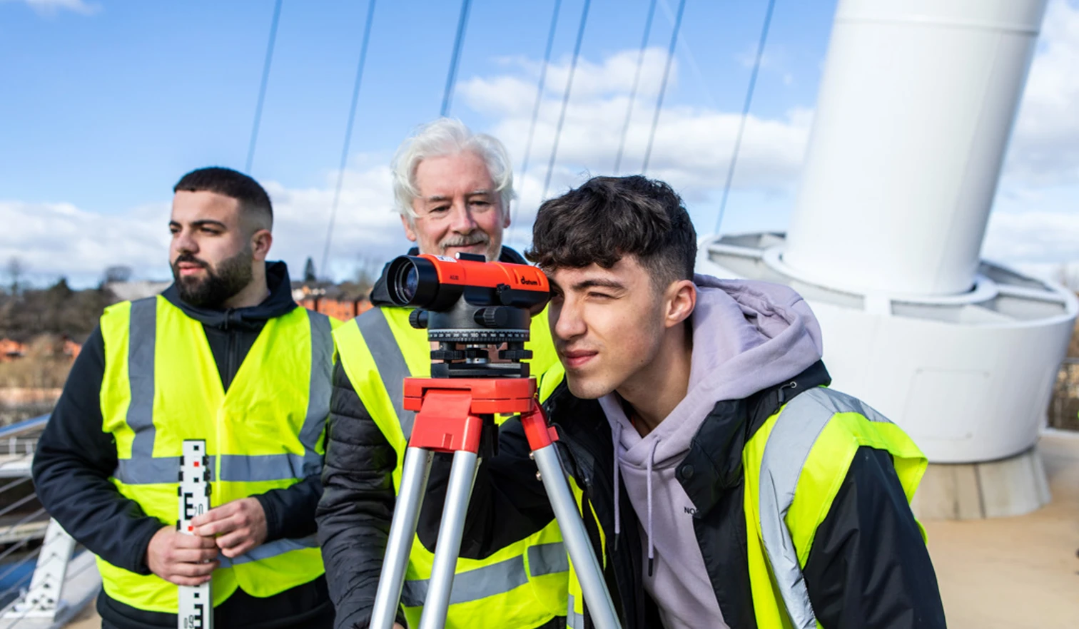 A young student in a high-visibility vest looking through a surveying instrument while the lecturer and another student observe and discuss measurements on a bridge A young student in a high-visibility vest looking through a surveying instrument while the lecturer and another student observe and discuss measurements on a bridge